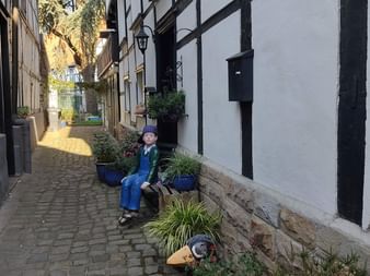 Kopfsteinpflastergasse in Hattingen gesäumt von weißen Fachwerkhäusern. Eine Figur sitzt auf einem Vorsprung neben Topfpflanzen entlang des schmalen Durchgangs.