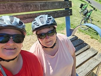 Zwei Radfahrer mit Helmen und Sonnenbrillen machen ein Selfie auf einer übergroßen Holzbank in Randersacker, Fahrräder stehen im Gras.