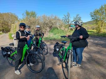 Four cyclists with green bikes and helmets stopped on a gravel path surrounded by trees and bushes under blue sky.