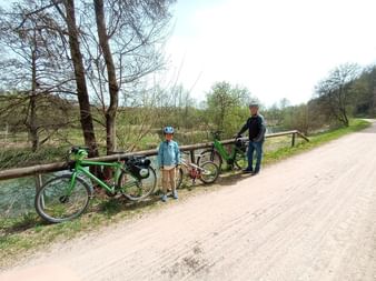 Erwachsener und Kind mit Fahrrädern neben Holzzaun am Vils-Radweg. Grünes Fahrrad im Vordergrund, Bäume und Bach im Hintergrund sichtbar.