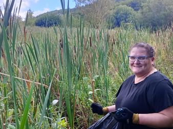 Woman named Katrin wearing glasses and black shirt, holding a garbage bag while standing in a wetland area with tall reeds and cattails in Burgwald.