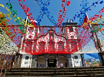 Weiße Kirche Nossa Senhora do Monte in Funchal mit zwei Glockentürmen, geschmückt mit bunten Papierblumen gegen blauen Himmel.
