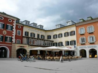 Hauptplatz in Lienz mit bunten historischen Gebäuden, Café-Außenbestuhlung unter weißen Markisen und geparkten Fahrrädern auf dem Platz.