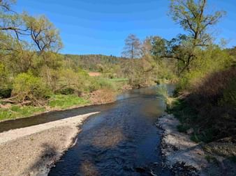 Shallow river with gravel banks flowing through green spring landscape with trees and hills under blue sky near Waldschlösschen.