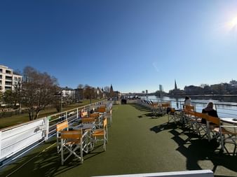 Sonnendeck mit Holzstühlen auf einem Flusskreuzfahrtschiff. Grüner Kunstrasen bedeckt das Deck. Stadtskyline und Fluss im Hintergrund unter blauem Himmel.