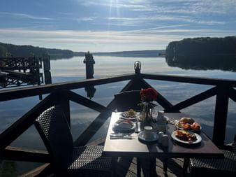 Frühstückstisch auf Holzterrasse mit Blick auf ruhigen See. Tisch gedeckt mit Kaffee, Gebäck und Blumen. Holzsteg und bewaldete Ufer sichtbar.