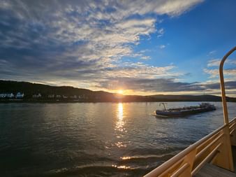 Sonnenuntergang über dem Rhein vom Bootsdeck aus, mit goldener Sonnenspiegelung auf dem Wasser, entferntem Passagierschiff und Hügeldorf.