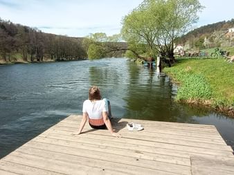 Person sitzt auf Holzsteg mit Blick auf Fluss in Pielenhofen. Grüne Hügel und Häuser im Hintergrund, Bootsverleih in der Nähe.