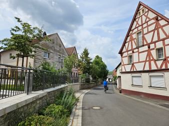 Radfahrer im blauen Shirt fährt durch Stammheim mit Fachwerkhaus und kleinem Bach entlang der Straße unter bewölktem Himmel.