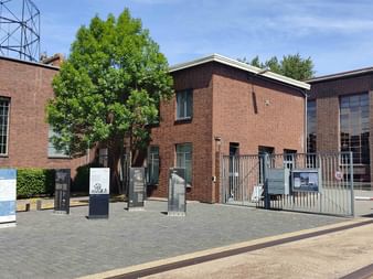 Eingangsbereich der Kokerei Hansa mit roten Backsteingebäuden, Metalltor, Informationstafeln und einem großen Baum. Links ist eine Metallturmstruktur sichtbar.