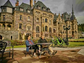 Two cyclists sitting at a table in front of an ornate historic castle with green bicycles parked nearby under a dramatic cloudy sky.