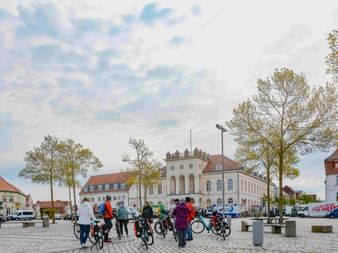 Gruppe von Radfahrern mit Fahrrädern auf einem Kopfsteinpflasterplatz vor historischen weißen Gebäuden mit roten Dächern unter bewölktem Himmel.