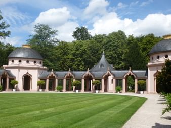 Pink baroque buildings with domed towers and arched openings in Schwetzingen Castle Park. Manicured lawn in foreground, dense forest backdrop.