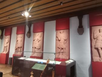 Museumsraum mit mittelalterlichen Steinreliefs an rot-weißen Wänden. Vitrinen mit Ausstellungsstücken im Vordergrund.