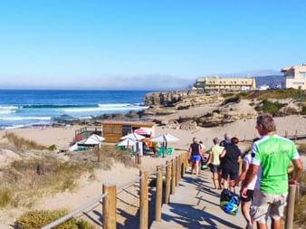 Cyclists walking to beach in Portugal Group of cyclists walking down wooden boardwalk toward Portuguese beach with blue ocean, rocky cliffs, and coastal buildings under clear sky.