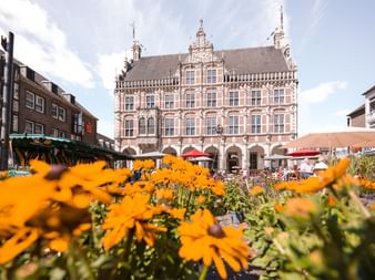 Orange Blumen im Vordergrund mit Bocholts prächtigem Renaissance-Rathaus dahinter. Marktstände und Cafés auf dem Platz unter blauem Himmel.