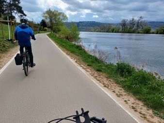 Radfahrer in blauer Jacke auf asphaltiertem Radweg entlang der Mosel mit grünen Hügeln und bewölktem Himmel im Hintergrund.