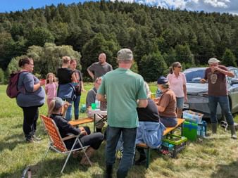 Group gathering outdoors in Burgwald with picnic benches and car. Dense forest hillside and blue sky with clouds in background.