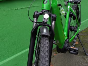 Green e-bike with illuminated front and rear lights parked against a green wall. The bike features a front dynamo light and rear carrier.