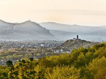 Panoramic view of Bonn with Drachenfels and Godesburg hills. Green vegetation in foreground, city buildings in valley, misty mountains in background.