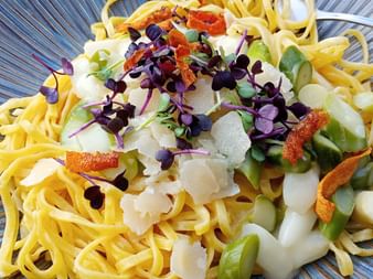 Gelbe Tagliatelle mit weißem Spargel, Parmesanspänen, lila Blüten und Kräutern auf einem blauen gerillten Teller.