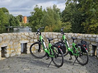 Zwei grüne E-Bikes mit schwarzen Taschen auf Kopfsteinpflaster an einer Steinbrückenmauer. Ein Fluss mit Bäumen und Gebäuden im Hintergrund.