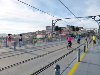 Radfahrer fahren auf der Dom Luís I Brücke mit Portos historischer Stadtsilhouette im Hintergrund. Bunte Gebäude säumen den Hang unter bewölktem Himmel.