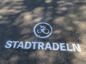 White STADTRADELN text and bicycle icon in circle painted on gray asphalt with dappled tree shadows.