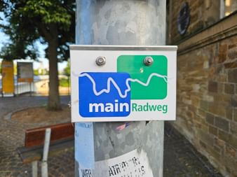 Weißes Schild mit blau-grünem Logo 'mainRadweg' an einem Metallmast auf einem Platz mit Backsteingebäuden und Bäumen.