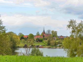 Dorf mit rotgedeckter Kirchturmspitze auf Hügel mit Blick auf ruhigen See, eingerahmt von grünen Bäumen. Traditionelle Häuser unter bewölktem Himmel.