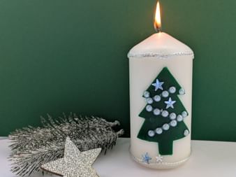 White pillar candle with lit flame, decorated with green Christmas tree, silver beads and stars. Silver star ornament beside it on white surface.