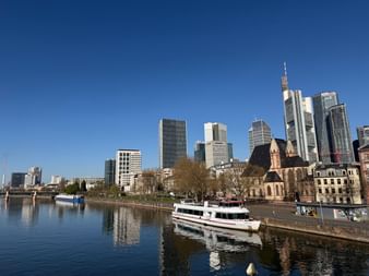 Frankfurter Skyline mit modernen Wolkenkratzern und historischer Kirche am Main. Ausflugsschiffe am Ufer unter klarem blauen Himmel.