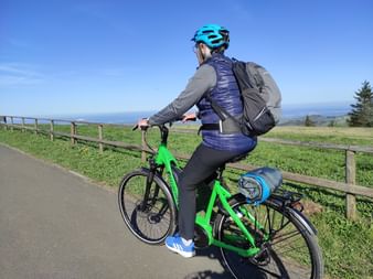 Cyclist with turquoise helmet on green e-bike along Fulda bike path, overlooking rolling hills and distant mountains under blue sky.