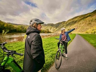 Two cyclists with helmets on a path by the Mosel River. One gestures toward terraced vineyards on hillsides under a cloudy sky.