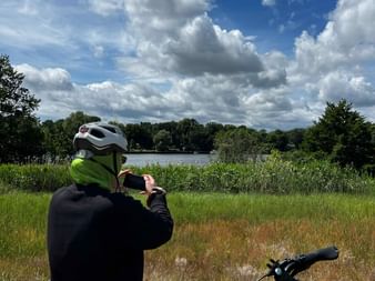 Radfahrer mit Helm fotografiert den Sacrower See. Fahrrad im Vordergrund, grüne Wiese und Wald unter blauem Himmel mit weißen Wolken.