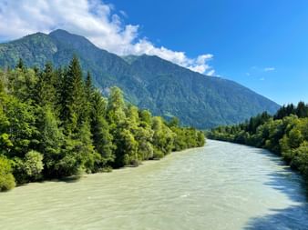 Türkisfarbene Drau fließt durch grünes bewaldetes Tal bei Steinfeld mit Bergen im Hintergrund unter blauem Himmel mit weißen Wolken.