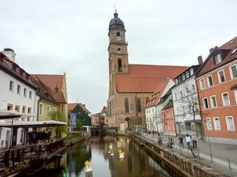 Kanalblick in Amberg an der Vils mit bunten Häusern, einer Kirche mit hohem Turm und Fachwerkhäusern, die sich im ruhigen Wasser spiegeln.