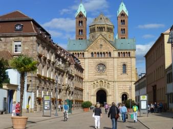 Speyer Cathedral facade with twin green-domed towers and Romanesque architecture. Visitors walk across the square with historic buildings on both sides.