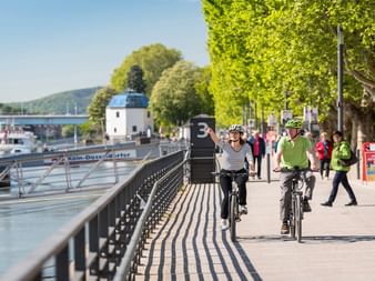 Cyclists on Koblenz riverside bike path Two cyclists riding along a paved riverside path in Koblenz with river boats, bridge, and green trees lining the waterfront promenade.