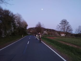 Group of cyclists riding on a paved country road at dusk with the moon visible in the sky. Trees and fields line the roadside.