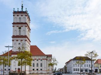Weiße Kirche mit hohem Turm und rotem Dach auf einem Marktplatz an der Havel. Der Platz ist von historischen Gebäuden und Bäumen umgeben.