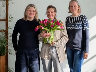 Three women standing together indoors. The woman in the center holds a large bouquet of pink and purple tulips, smiling with colleagues.