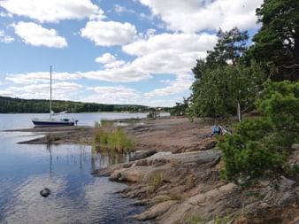 Felsige Küste im Naturpark Erstavik Felsige Küste im Naturpark Erstavik mit Segelboot im ruhigen Wasser. Kiefern säumen die Granitküste unter bewölktem blauen Himmel.