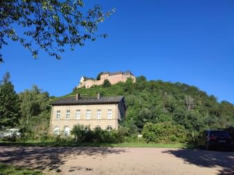 Burg auf einem weinbewachsenen Hügel über einem beigen zweistöckigen Gebäude mit Bogenfenstern. Klarer blauer Himmel und Bäume rahmen die Szene.