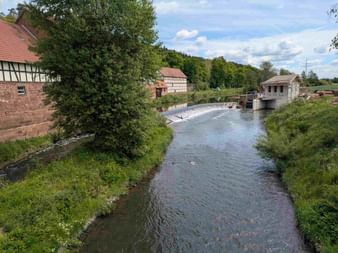 River flowing through Hessen with a weir, half-timbered houses on the left, a small hydroelectric station on the right, green banks and trees.
