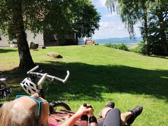 Two cyclists relaxing on wooden benches with drinks and a bicycle nearby, overlooking a green lawn with trees and distant hills under blue sky.