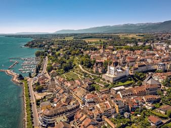 Aerial view of Nyon, a historic town on Lake Geneva with terracotta roofs, a castle, marina, and mountains in the background.