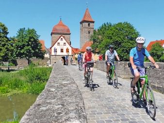 Cyclists on historic stone bridge in Germany Three cyclists riding across a medieval stone bridge with cobblestones. Behind them stands a historic town with towers and red roofs.
