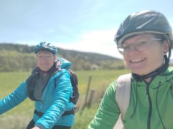 Zwei lächelnde Radfahrer mit Helmen und Brillen machen ein Selfie auf dem Fulda-Radweg, mit grünen Feldern und Hügeln im Hintergrund.