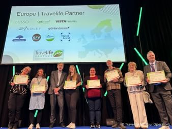 Group of eight people holding Travelife sustainability certificates on stage at ITB. Screen displays Europe Travelife Partner logos.